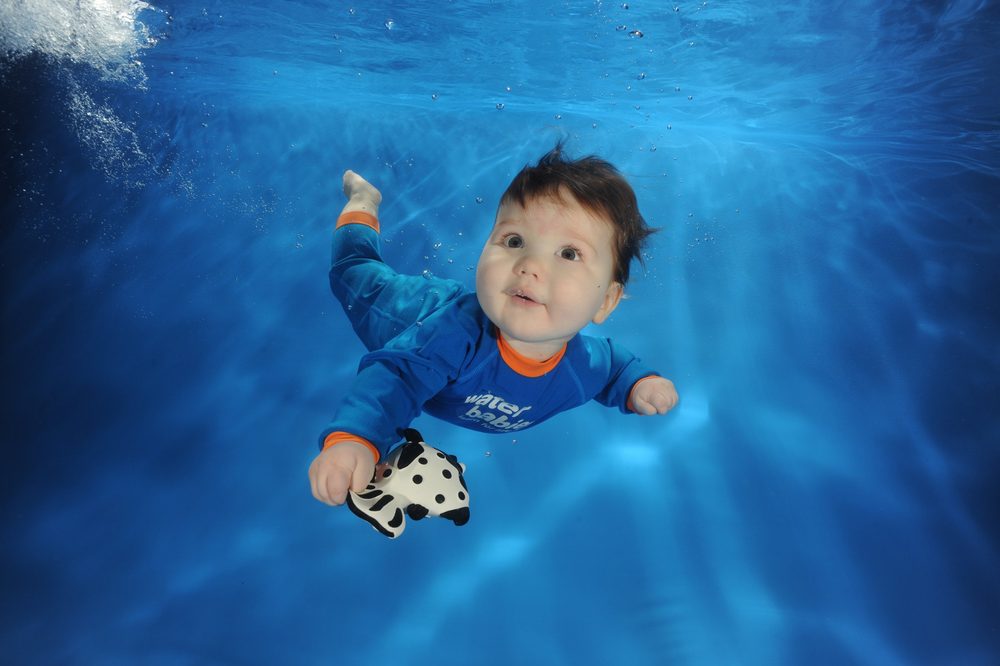 A smiling baby swimming underwater in a Water Babies suit, holding a toy zebra